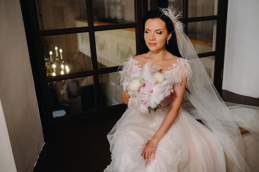 A bride in a wedding dress and a bouquet sits at an open old window and looks.