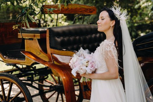 A stylish bride with a bouquet stands near a carriage in nature in retro style.