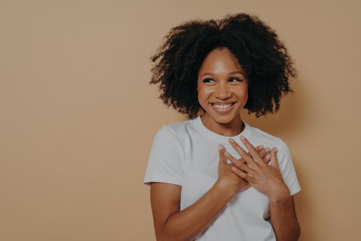 Portrait of happy african woman in white t-shirt posing over beige background with smile looking away, holding hands on chest feels gratitude, gesture of sincere feelings from heart and love concept