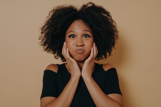 Cute african woman with wavy hairstyle making funny face, puffs out her cheeks and holds them with hands. Indoor portrait of glamorous african model isolated on dark beige background with copy space