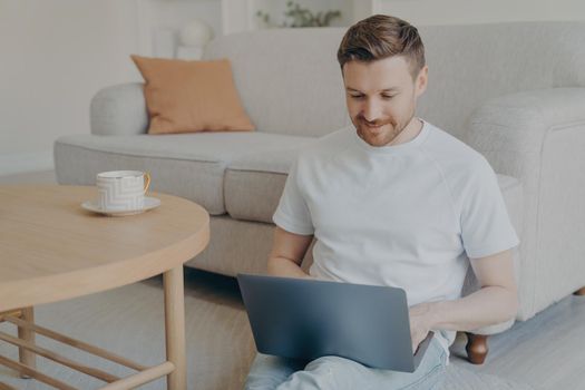 Attractive caucasian bearded freelancer man having his morning cup of coffee while sitting on carpeted floor in modern living room interior with laptop, surfing internet and working online from home