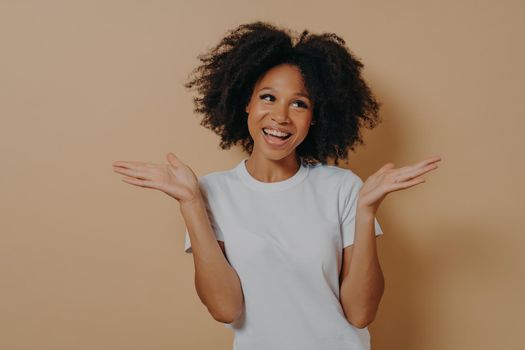 Young smiling mixed race curly woman raising both hands with hesitation and looking aside, making decision or choosing between something, dressed in white tshirt, isolated on beige background