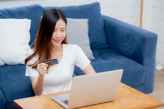 Young asian woman sit on sofa using laptop computer shopping online with credit card buying to internet, happy girl payment with e-business on couch, purchase and payment, business concept.