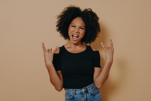 Rock star. Young self confident afro american woman holding both hands in rock n roll sign and shouting loudly with emotional face expression, feeling carefree while posing against beige background