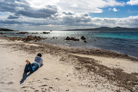 Young single woman looking at awesome cloudy sky lying on the beach sand in autumn or winter season thinking about future destiny. Lonely girl in contact with nature to discover itself