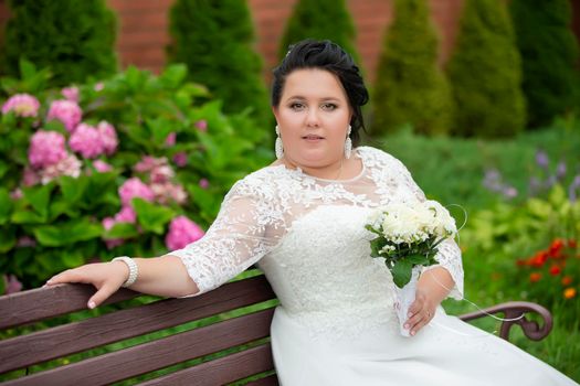 Portrait of a plump bride with a bouquet, who is sitting on a bench and looking at the camera.