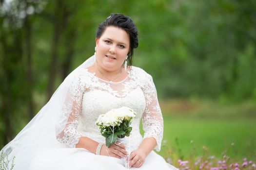 Portrait of a full bride with a white wedding bouquet on a forest background.