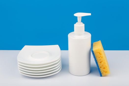 Dishwashing concept, creative composition with liquid detergent in white plastic bottle with dispenser, yellow cleaning sponge and pile of clean plates. High angle view.