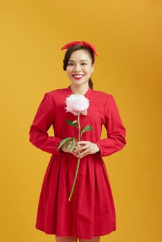 Portrait of adorable young woman standing with charming smile, holding peony flower in hands and smells it.