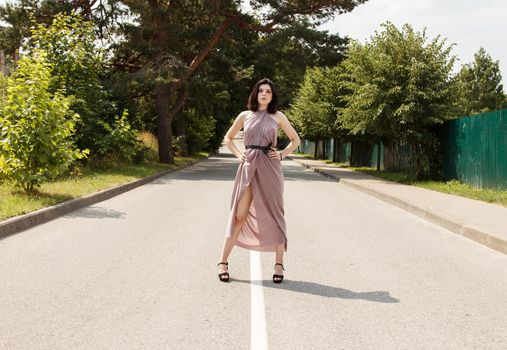 young beautiful brunette woman in beige dress standing on the road in summer
