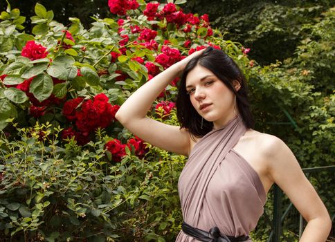 portrait of a young brunette woman standing by a rose bush on summer day. outdoor closeup