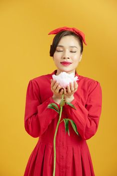 Portrait of adorable young woman standing with charming smile, holding peony flower in hands and smells it.