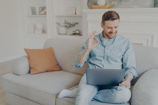 Handsome caucasian male freelancer smiling and showing sign OK with fingers while having video conference on notebook, young man in casual clothes using wireless laptop for online meeting from home