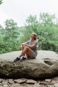 Travel concept. Young woman in summer dress sitting on a big rock in the forest, having rest or meditating