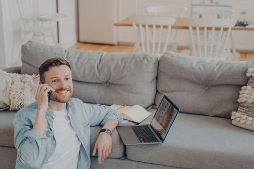 Young male office worker working from home, talking on phone with his co-worker about projects status, preparing presentation with gathered data, resting hand on couch while sitting on floor