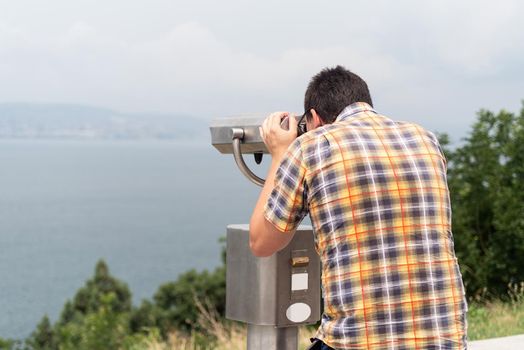 a young man in a colorful shirt watching what is happening with stationary binoculars on a warm summer day on a hill against the background of the mountains