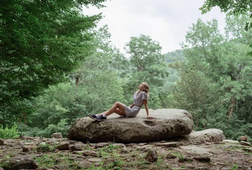 Travel concept. Young woman in summer dress sitting on a big rock in the forest, having rest or meditating