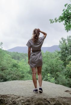 Travel and freedom concept. Back view of a young woman in summer dress standing on a big rock in the forest looking away