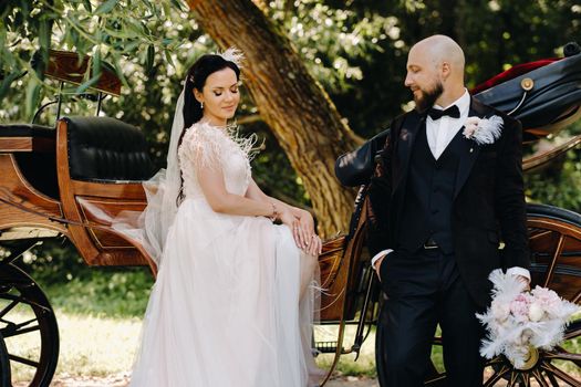 A couple of the bride and groom are standing near the carriage in nature in retro style.