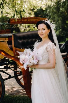 A stylish bride with a bouquet stands near a carriage in nature in retro style.