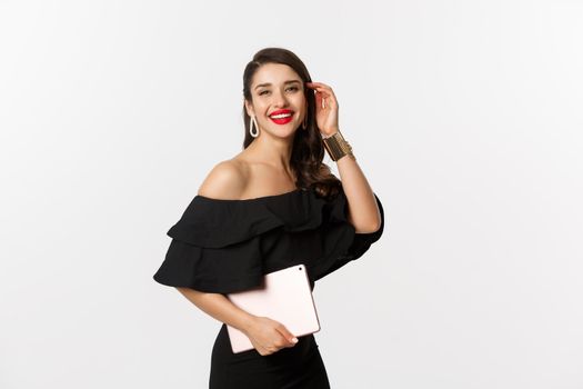 Fashion and shopping concept. Stylish young woman with glamour makeup, wearing black dress, holding digital tablet and smiling, white background.