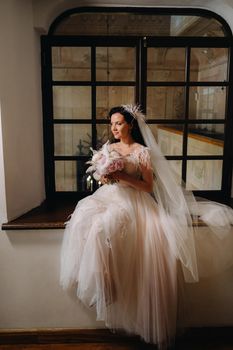 A bride in a wedding dress and a bouquet sits at an open old window and looks.