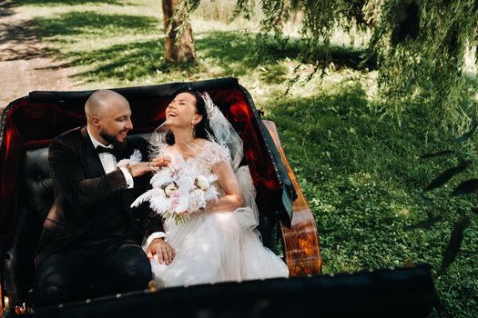 The bride and groom with a bouquet are sitting in a carriage in nature in retro style.