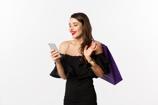 Beauty and shopping concept. Gorgeous woman in elegant black dress and makeup, order online on smartphone, holding bag and smiling, standing over white background.