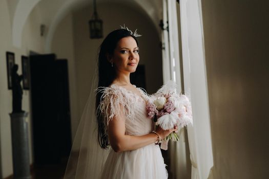 The bride in a wedding dress and with a bouquet stands at the old window and looks.