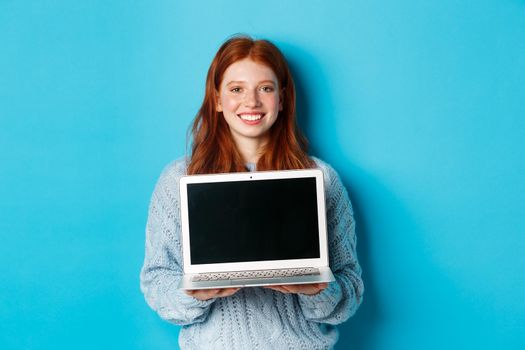 Young smiling woman with red hair and freckles showing computer screen, holding laptop and demonstrate online promo, standing over blue background.