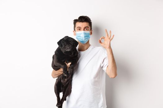 Covid-19, animals and quarantine concept. Young man in medical mask holding cute black pug dog, showing okay sign, like and approve, standing over white background.