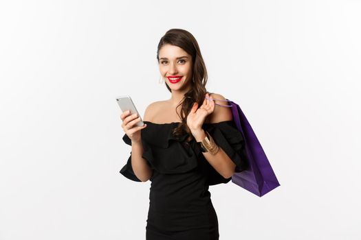 Beauty and shopping concept. Gorgeous woman in elegant black dress and makeup, order online on mobile phone, holding bag and smiling, standing over white background.