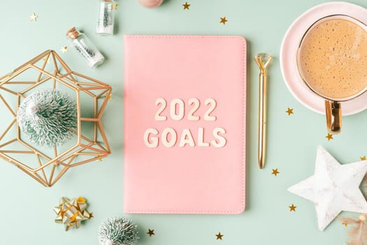 Top view Desktop Christmas pink notepad with 2022 goals letters text. Flat lay on green mint table background with planner, cup of coffee, candle, Christmas decoration, notebook and stationery.