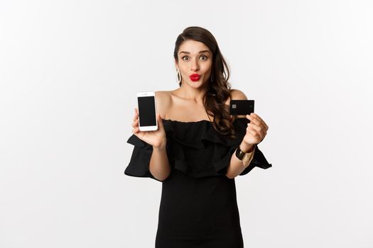 Fashion and online shopping concept. Happy young woman in black dress, showing credit card and mobile screen, standing over white background.