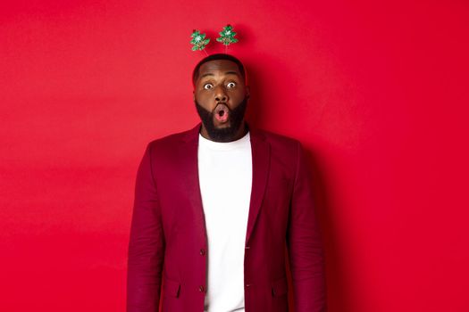 Merry Christmas. Happy african american man celebarting New Year, wearing party headband and making funny faces , standing over red background.