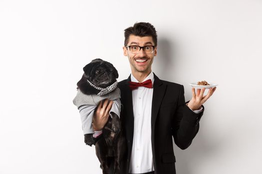 Handsome young hipster in suit and glasses holding cute black pug and pet food on plate, standing over white background.