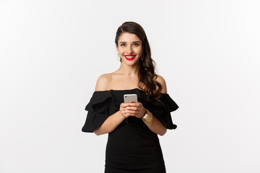 Online shopping concept. Attractive young woman in black dress, reading text message, using mobile phone and smiling, standing over white background.