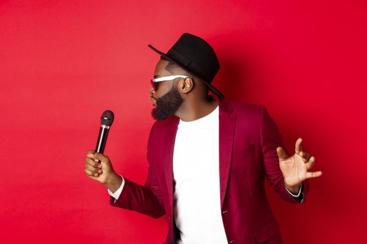 Passionate black male singer performing against red background, singing into microphone, wearing party outfit, standing over red background.