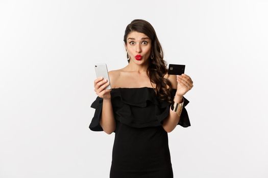 Online shopping concept. Fashionable woman in black dress, holding credit card with smartphone, looking excited, standing over white background.
