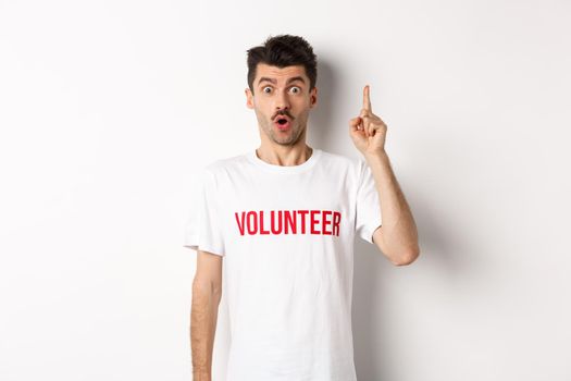 Handsome young man in volunteer t-shirt having an idea, raising finger and saying suggestion, pointing up, standing over white background.