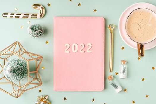 Top view Desktop Christmas pink notepad with 2022 letters text. Flat lay on green mint table background with planner, cup of coffee, candle, Christmas decoration, notebook and stationery.