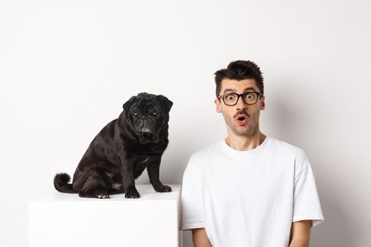 Portrait of dog owner and small cute pug staring at camera surprised and amazed, standing over white background.