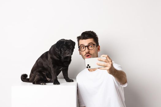 Image of handsome young man taking selfie with cute black dog on smartphone, posing with pug over white background.