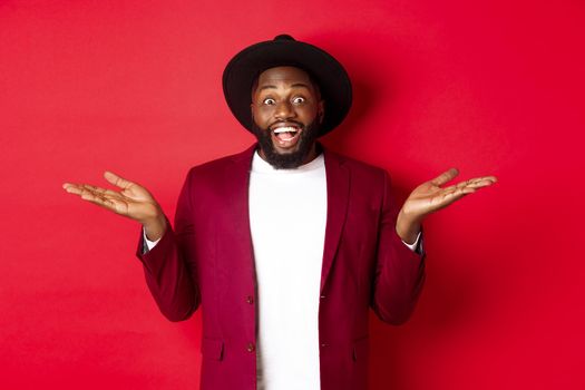 Christmas shopping and people concept. Handsome african american man smiling, spread hands sideways, showing promo offers on copy space, red background.