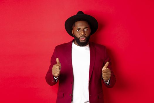 Impressed Black man showing thumbs up and staring amazed at camera, approve and praise something good, red background.