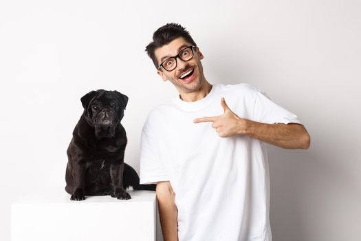 Happy young man showing his cute dog, pointing finger at black pug and smiling, standing over white background.