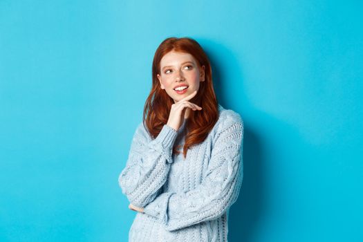Thoughtful teenage girl with red hair, looking upper right corner logo and thinking, imaging something, standing over blue background.
