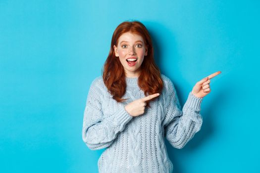 Winter holidays and people concept. Curious teenage girl in sweater, pointing fingers right and staring at camera amazed, showing promo offer, standing over blue background.