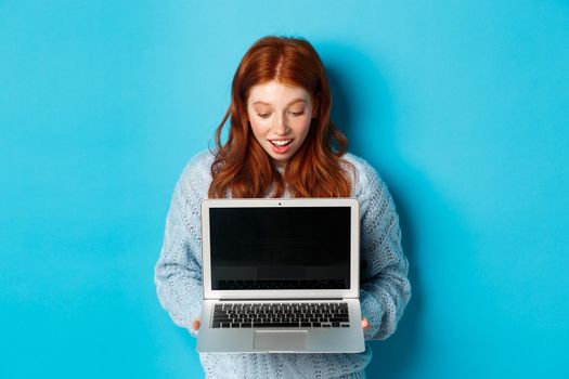 Amazed redhead girl staring at laptop screen and looking impressed, showing computer display, standing over blue background.