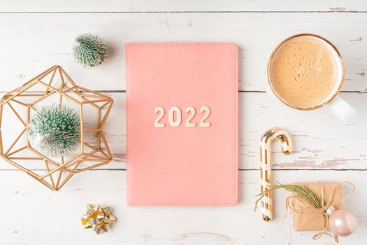 Top view Desktop Christmas pink notepad with 2022 letters text. Flat lay on white wooden table background with planner, cup of coffee, candle, toys, Christmas decoration, notebook and stationery.
